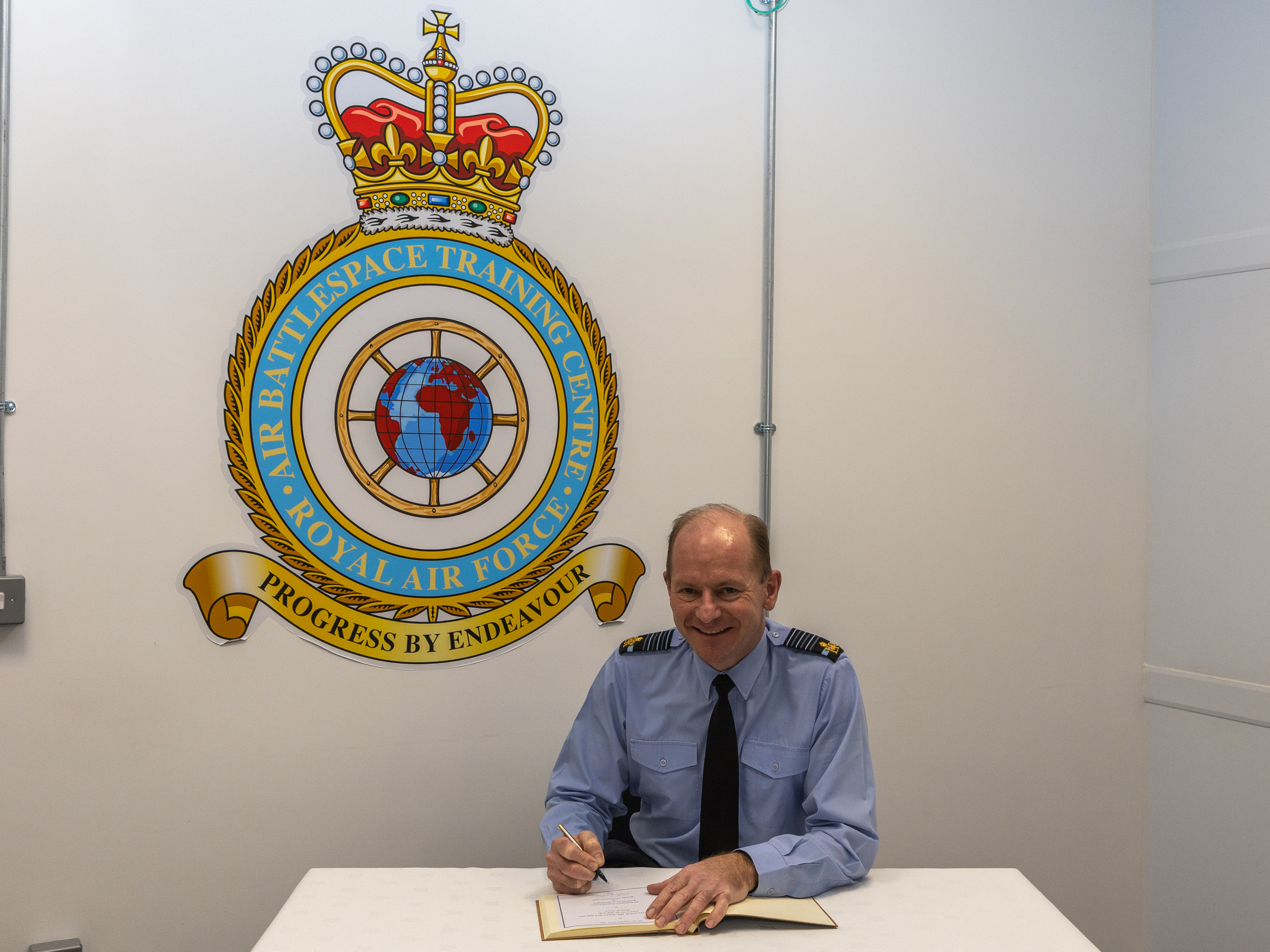 Image show the Chief of the Air Staff sat at a table signing a book, with the Air Battlespace Training Centre badge on the wall.